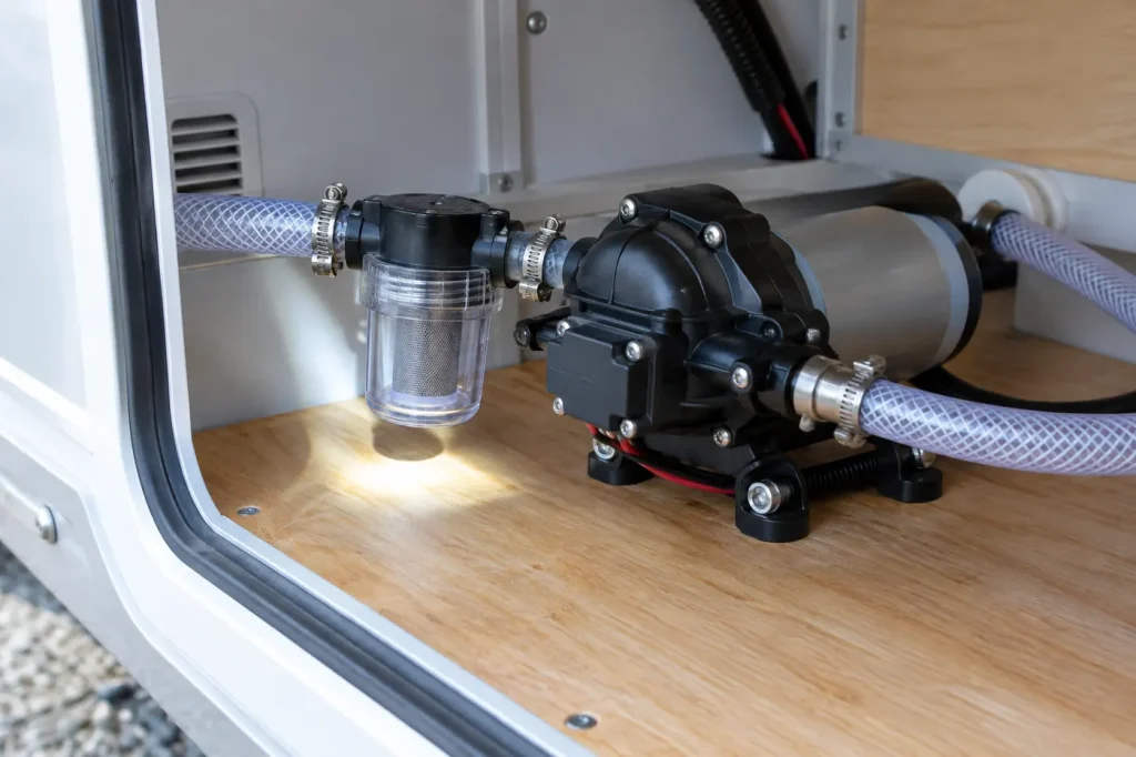RV water pump with clear inlet strainer bowl and hoses in a storage bay
